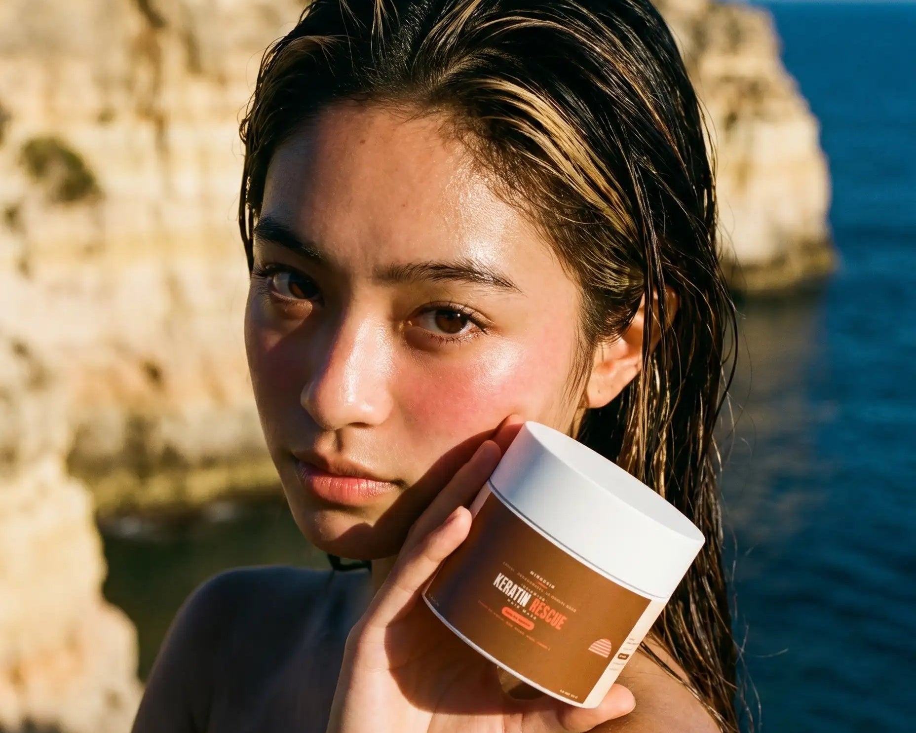 Woman holding a jar of Keratin Mask against a scenic coastal background