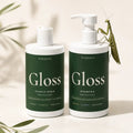 Two MirenSkin Gloss haircare bottles on a warm cream background with botanical props. One bottle is clear with a white flip-top cap and the other is a white pump bottle, both with dark green labels reading Gloss Conditioner and Gloss Shampoo.